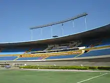 Lateral section of an oval shaped stadium, with empty blue and white seatings below floodlights, viewed from the pitch; the sky is blue and cloudless.