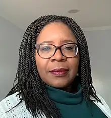 A Black woman with long braids and glasses. Her subtly made up face shows quiet confidence