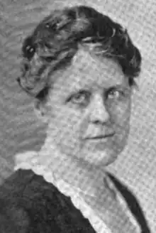 A black-and-white image of Esther Dunshee, a white woman, wearing a dark dress or blouse with a white lace collar; her eyes appear light in color, and she has pale or absent eyebrows