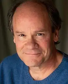 A white man with greying brown hair and a blue sweater is smiling into the camera.