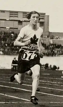 A woman sprints down a track during a race. She has short hair and is wearing dark shorts and a white shirt with a stylized maple leaf logo over the word "CANADA".