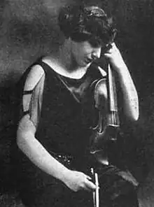 A white woman seated, holding a violin, looking downward, wearing a black dress with bare shoulders. Her hair is short or in an updo, off her neck.