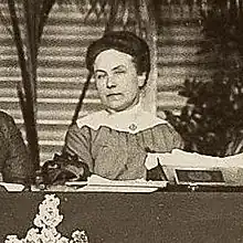 A woman in a dark dress with a wide white collar sitting behind a dais which has papers on it and horizontal striped panels and plants in back of her.