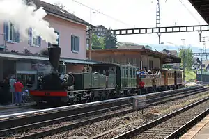 Green steam locomotive with carriages in front of a two-story building