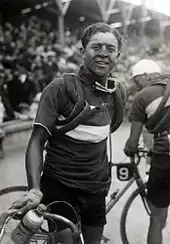 A man with a dirty face holding his bicycle, spare tires wrapped around his shoulders. In the background a large crowd.