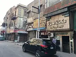 A street with parked cars and several shops with appartements above them.