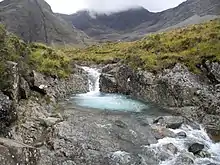 In a wild mountainous landscape, a small waterfall leads into a pool with a striking cyan colour