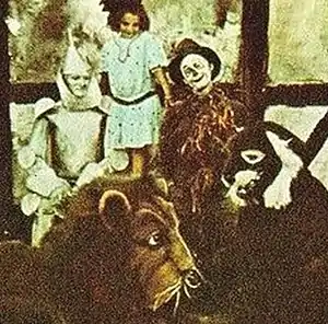 A colour-tinted monochrome photograph of a group of actors. At the rear, a young girl. In front of her, the iron man and scarecrow from 'The Wizard of Oz'; in front of them, people dressed as a lion and another animal.