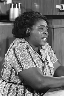 An African American woman sits at a conference, and appears to be speaking. Her hair is short and she is wearing a dress with a kaleidoscopic pattern. The picture is in black and white.