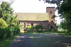 A photograph of a simple country church, with its square western tower in brick to the right and the older stone eastern end, slightly obscured by trees to the left.