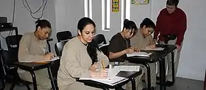 Four female prisoners in beige uniforms seated at desks . A teacher is supervising one of them.