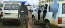 A soldier with a blue cap between two white cars. The car on the right holds a United Nations flag