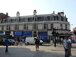 A white building with a blue-shingled roof and a blue sign reading "FINCHLEY ROAD STATION" in white letters all under a blue sky with white clouds