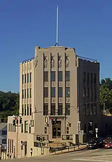 A four-story tall light brown stone building with a flagpole on top. Its sides flare out in the rear following the streets on either side, and there is decorative carving in the stone at the top