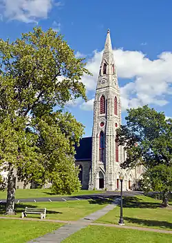 A tall pointed light brown stone spire with red trim and narrow lancet windows and vents, attached to a similar church building, rises between two oak trees in a parklike setting under blue skies