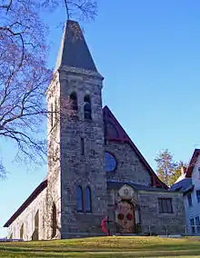 A triangular stone building with a peaked-roofed tower attached to its front slightly to the left, illuminated by the sun from that direction. Its wooden doors have a Christmas wreath on them.