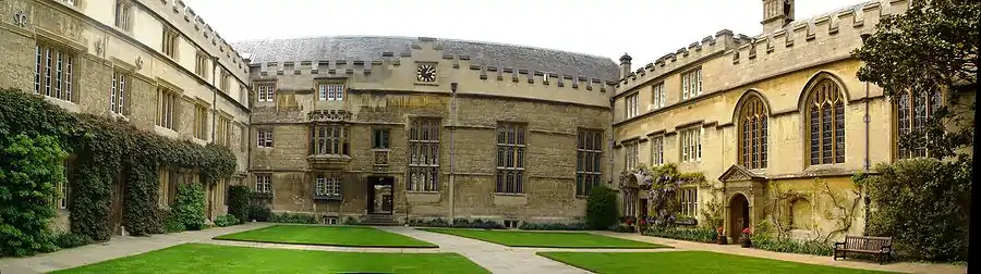 Adjoining buildings (all with battlements) on three sides of an area of grass in the middle of a quadrangle crossed by pathways from side to side and front to back; at the far side, a large building with a clock at roof level and three large windows; on the left, regularly placed windows in a three-storey building; on the right, a chapel with three arched windows alongside a three-storey house