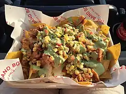 A cardboard tray lined with white paper reading "The Band Box – Right Field" holds yellow corn chips, smoked chicken pieces, jalapeño queso, sweet yellow corn, pico de gallo, and green cilantro crema.