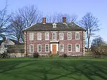 A two-storey brick-built mansion in its own well-kept garden of green grass. The ground floor has six windows – three on each side of a wooden front door. The second floor has seven windows. The house has a grey slate roof surmounted by two small brick chimneys. Behind the house are leafless trees and behind those, a clear blue sky.