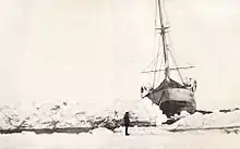 Front view of ship showing it listing to the right. It is surrounded by ice, but in front of the ship a narrow channel of open water is visible. The scene is watched by a man standing on the ice.