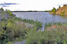 A different view of the lake with a shallow weedy area in the foreground, and a high area of mostly bare rock at the upper right