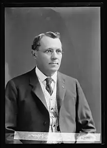 A man with wavy, graying hair wearing glasses, a white shirt, light vest, and a dark tie and jacket