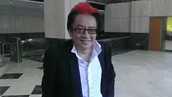 Frankie Kao wearing a white ruffled shirt and dark suit jacket, with a red blaze in his hair, grinning at camera