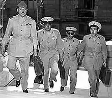 Four men in military uniform, wearing buttoned jackets, wide-rimmed hats, black shoes, and carrying briefcases. They are ascending the stairs of a building.