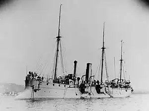 A ship with three tall masts and two smoke stacks lies anchored in shallow water offshore