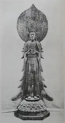 Front view of a standing statue with flowing robes holding a small container in front of her body. There is a large halo behind the head of the statue.