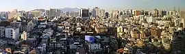 A daytime high-angle shot of a number of older mid-sized commercial and residential buildings on an incline. In the background, some larger office, department store, and church buildings are visible. In the very back, mountains and the North Seoul Tower are visible.