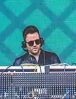 A dark-haired man performs on a set of turntables