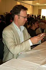 A man facing the right focused on his upper body. He is wearing a light brown jacket, and a white shirt.