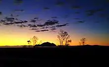 Small, pointed mountain on the horizon, foregrounded by scattered bare trees, silhouetted against the sky at sunset