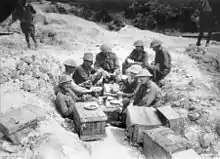 Eight men, most wearing steel helmets, sit in a shell hole surrounded by wooden crates.