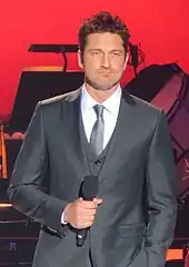 Gerard Butler standing with a microphone with a red background in a suit and tie facing the camera