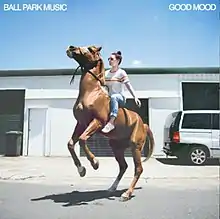 A woman atop a rearing horse in front of the band's studio, on a sunny day.