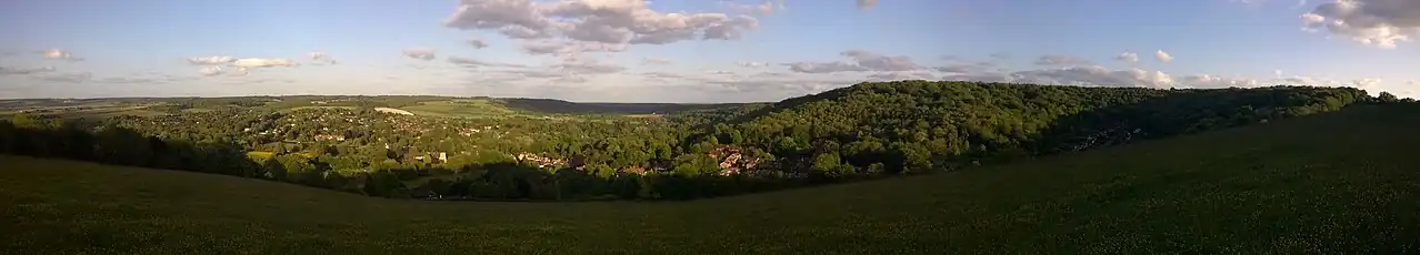 View over tree-covered hills on a sunny evening. Houses of a large settlement are visible between the trees.