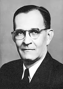 Black and white photograph of a white man with dark hair, wearing a suit and glasses