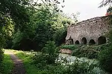 A footpath to the left, water in the centre of the picture and on the right a stone wall with arch shaped openings.