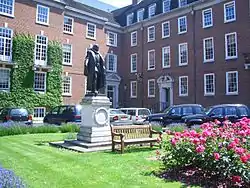 A grassy area with floral shrubs and a central statue on a plinth, with terraced buildings in the background