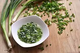 A small bowl of oil with green onion in it. This mix has been well heat in a pan. Nearby are whole green onions and a few chopped green onion. It looks like they are on a brown, wooden chopping board