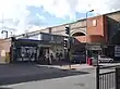 A red-bricked building with a blue sign reading "GREENFORD STATION" in white letters and people in front all under a blue sky with white clouds