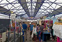 An interior of a building with a translucent glass roof supported by blue-painted steel latticework. On the main floor are a number of different stalls with customers inspecting various wares.