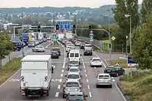 Vehicles backed up on a divided roadway seen from above. In the foreground is a traffic signal; there are blue signs in German further down the road as it narrows. In the rear is a developed hillside, partially obscured by bluish haze