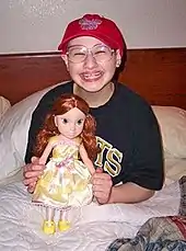 A young woman sitting upright in a bed, her legs under the blanket and the headboard and pillows behind her. She holds a large doll in front of her and is wearing a T-shirt, large glasses and a red baseball cap while smiling broadly