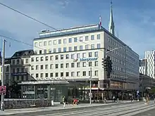 A 7 story concrete building stands in a street in winter. The building is pale and unadorned. Small windows form rows on each floor. The bottom level has commercial spaces and a small transit station and a church steeple are also in frame.