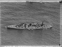 An aerial photograph of a large, World War I-era warship armed with four gun turrets, each with two guns, at sea. The ship is listing to port, but does not appear to be otherwise moving.