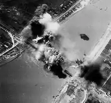 A black-and-white photograph showing an explosion on a railway bridge, with an American bomber flying overhead.