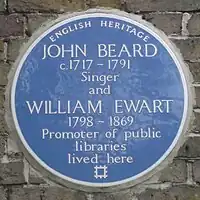 A Blue plaque on a brick wall with the words "John Beard C1717 - 1791 Singer and William Ewart 1798 - 1861 Promoter of Public Libraries